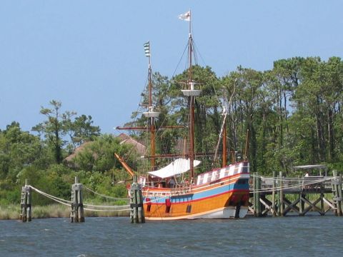 1985 Ford LTD Crown Victoria - A historic sailing ship is moored to a pier with trees in the background.