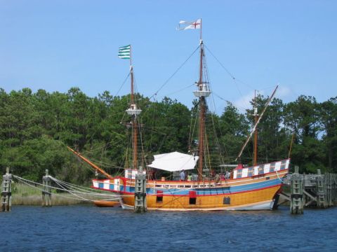 1985 Ford LTD Crown Victoria - A historic sailing ship is moored at a dock, with trees lining the shore under a clear blue sky.