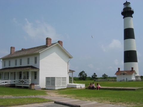 1985 Ford LTD Crown Victoria - A lighthouse and adjacent keeper's house stand under a partly cloudy sky, with a few people relaxing on the lawn.