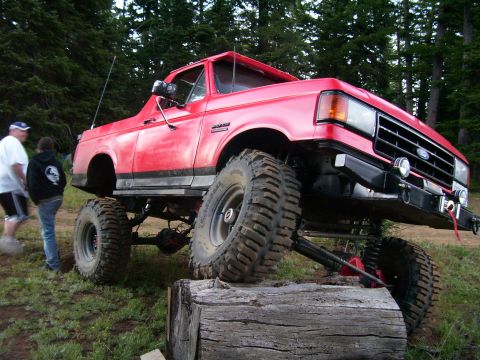 1989 Ford Bronco - A red 1989 Ford Bronco is shown with its suspension lifted, positioned on a large log during an off-road event.