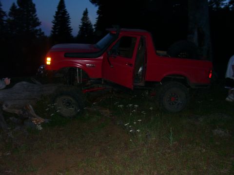 1989 Ford Bronco - A red Ford Bronco with its driver door ajar sits on uneven terrain as twilight descends.