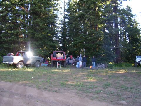 1989 Ford Bronco - A group of people gather around a campfire near several parked vehicles in a wooded area.