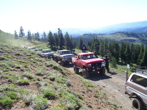 1989 Ford Bronco - Several modified trucks drive up a steep, dusty mountain road with a scenic overlook.