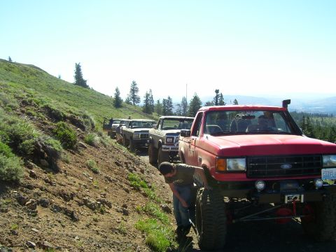 1989 Ford Bronco - A line of modified Ford Broncos navigate a mountain trail under a clear blue sky.