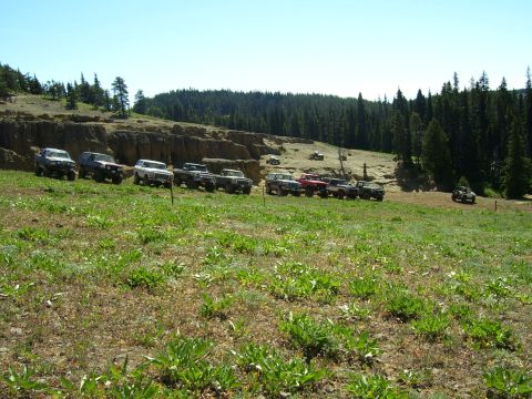 1989 Ford Bronco - Several off-road vehicles are gathered on a grassy hillside with a wooded area and cliffs in the distance.