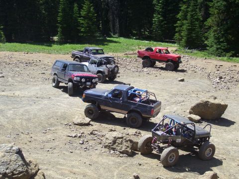 1989 Ford Bronco - A group of modified off-road vehicles, including a 1989 Ford Bronco, navigate a rocky trail in a wooded area.