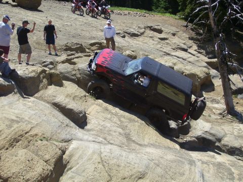 1989 Ford Bronco - A black 1989 Ford Bronco with flame decals navigates a steep, rocky trail while onlookers watch.