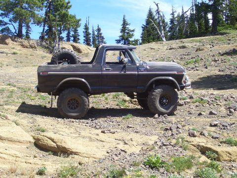 1989 Ford Bronco - A lifted Ford Bronco sits on a rocky trail under a clear blue sky, surrounded by sparse vegetation and trees.