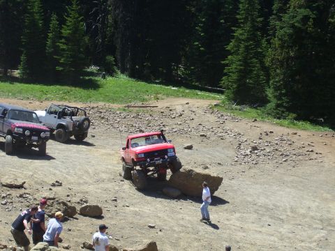 1989 Ford Bronco - A red pickup truck maneuvers over a large rock on a dirt trail, with other off-road vehicles visible in the background.