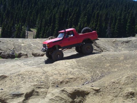 1989 Ford Bronco - A red off-road truck navigates a challenging dirt terrain with a forest in the background.