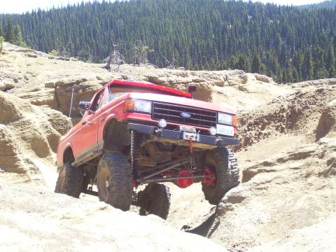 1989 Ford Bronco - This modified Ford Bronco navigates a steep, sandy incline with a dense forest in the background.