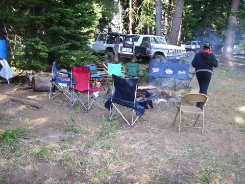 1989 Ford Bronco - A campsite scene with a fire pit, camping chairs, and two off-road vehicles parked in the background.