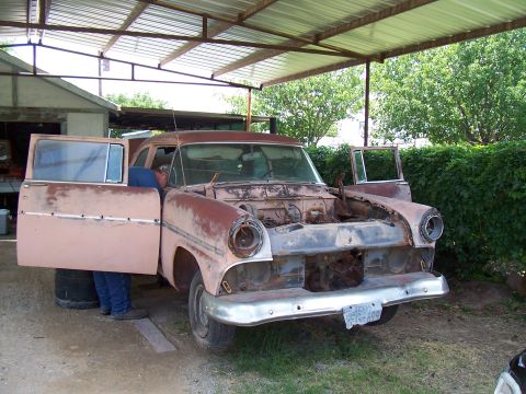 1955 Ford Customline - A rusty 1955 Ford Customline sits under a carport with its hood and doors open, revealing its disassembled front end.