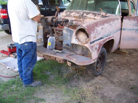 1955 Ford Customline - A person works on the front end of a 1955 Ford Customline, which is missing its headlight and has its engine exposed.