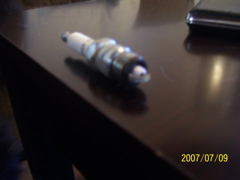 1990 Jeep Cherokee - A single spark plug is shown on a dark, reflective surface, with a date and website visible.