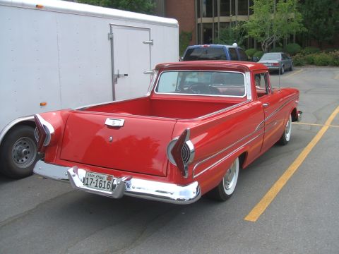 1974 Ford Ranchero - A red 1957 Ford Ranchero is parked next to a white trailer, with other vehicles and a building in the background.