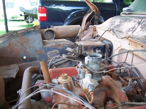 1955 Ford Customline - The engine bay of a weathered vehicle is visible, with a blue pickup truck and boat trailer in the background.