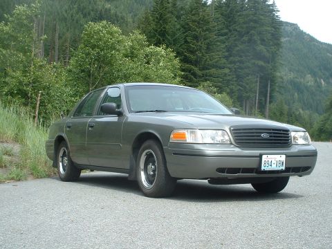 2000 Ford Crown Victoria - A gray 2000 Ford Crown Victoria is parked on the side of a road with a wooded mountainside behind it.