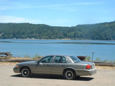 2000 Ford Crown Victoria - A gray 2000 Ford Crown Victoria is parked near a large body of water with a tree-covered hill in the background.