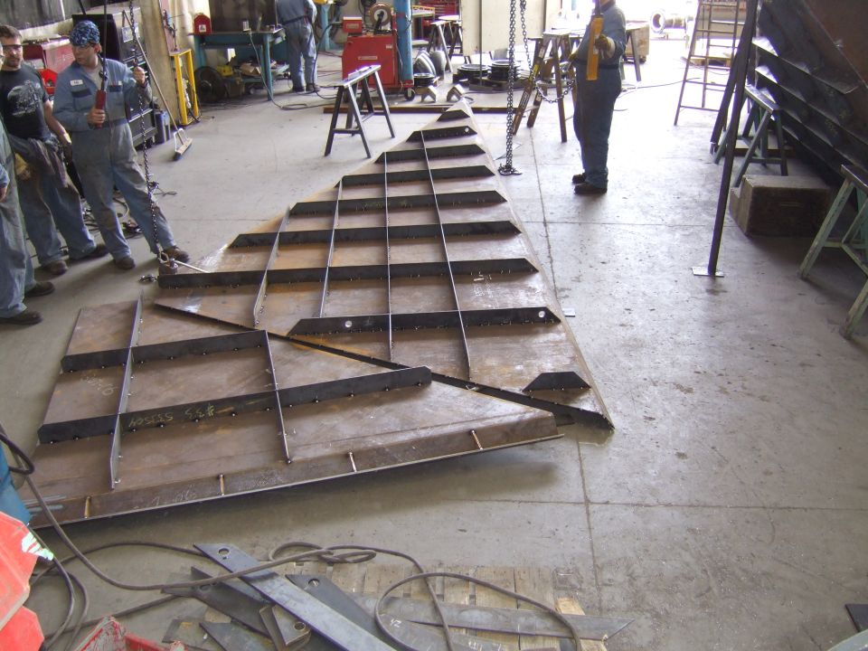 Large metal structure with grid of supports on concrete floor, workers nearby