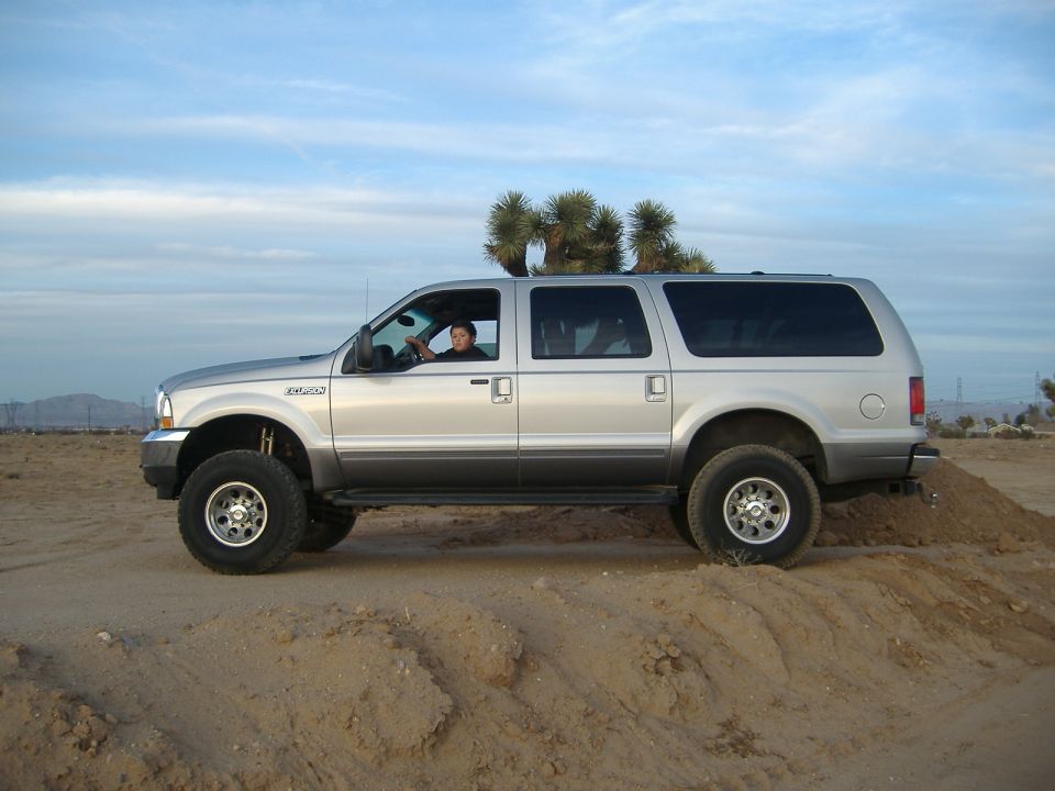 Silver lifted Ford Excursion SUV in side profile on desert dirt, Joshua trees behind, driver visible in window