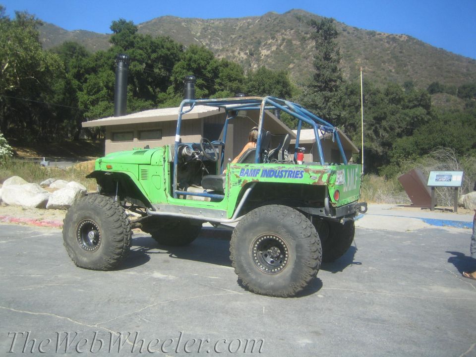 Green off-road vehicle with roll cage and big tires parked near a building and mountains