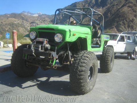 1987 Suzuki Samurai - A heavily modified green off-road vehicle with a roll cage is parked next to a white SUV.