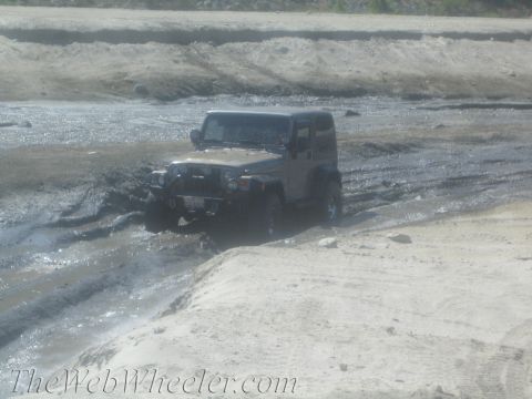 1987 Suzuki Samurai - A tan Jeep Wrangler navigates a muddy terrain, kicking up water as it drives through the mire.