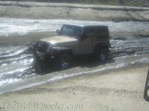 1987 Suzuki Samurai - A Jeep Wrangler navigates through a muddy, watery terrain, with its front right wheel deep in the mire.