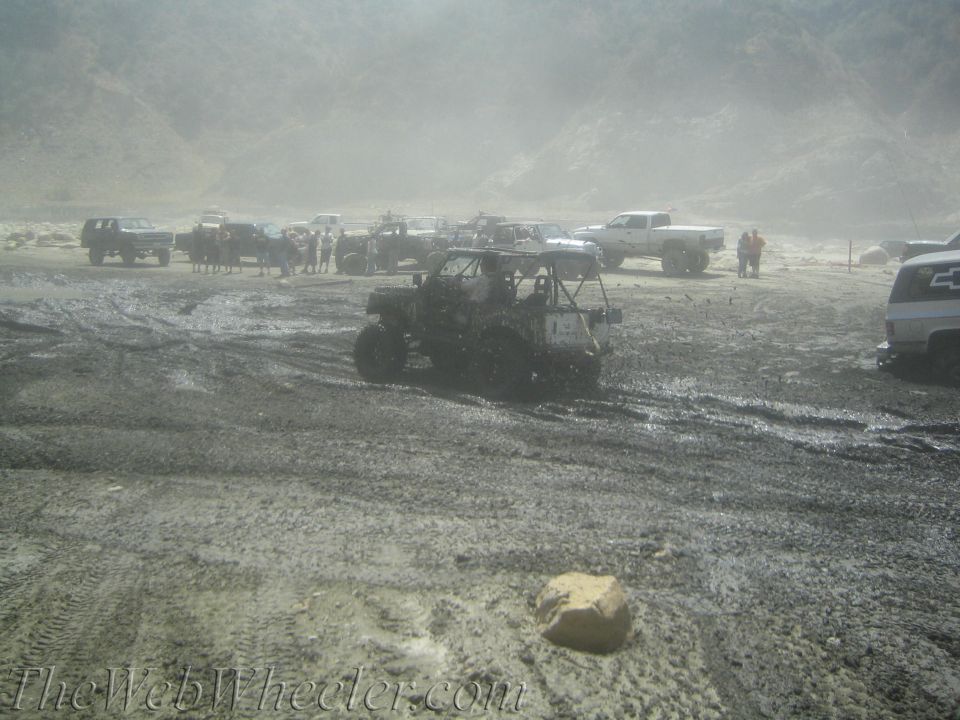 Muddy off-road vehicles gathered in a dusty, hilly area with tire tracks visible in the dirt