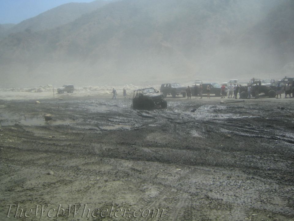 Several off-road vehicles are stuck in thick mud with people standing around. Mountains are in the background