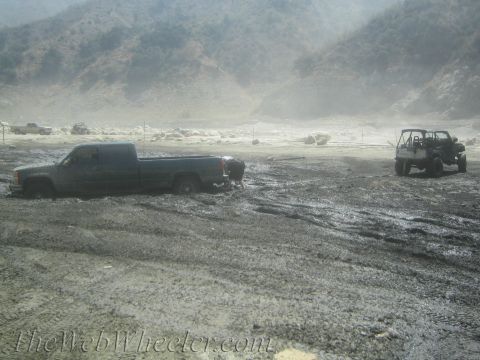 1987 Suzuki Samurai - A dark pickup truck is stuck in a muddy, off-road environment with another vehicle visible in the distance.