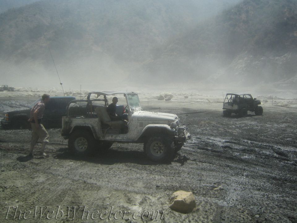 Several off-road vehicles are stuck in mud with people around them. A white Jeep-like vehicle is in the foreground