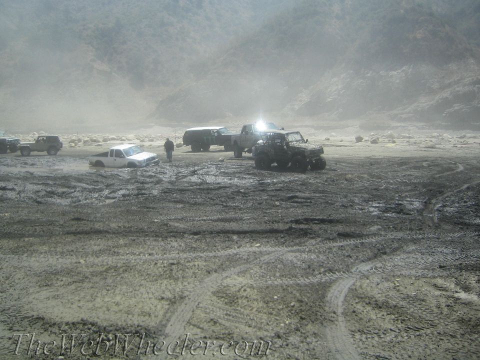 Muddy scene with several off-road vehicles, one white pickup truck stuck in mud