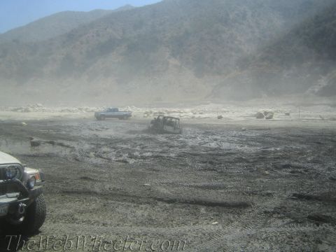 1987 Suzuki Samurai - Several off-road vehicles navigate a muddy, dusty terrain with mountains in the background.
