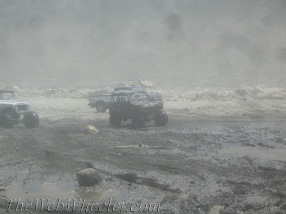 Three off-road trucks navigate a muddy, dusty landscape