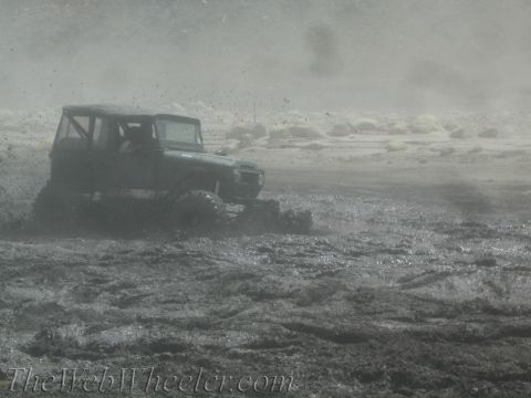 1987 Suzuki Samurai - A dark-colored off-road vehicle navigates through a muddy terrain, kicking up spray.