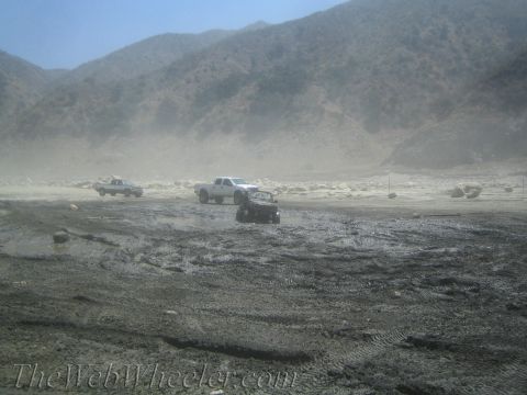 1987 Suzuki Samurai - Multiple off-road vehicles traverse a challenging muddy and dusty landscape under a clear sky, bright sky.