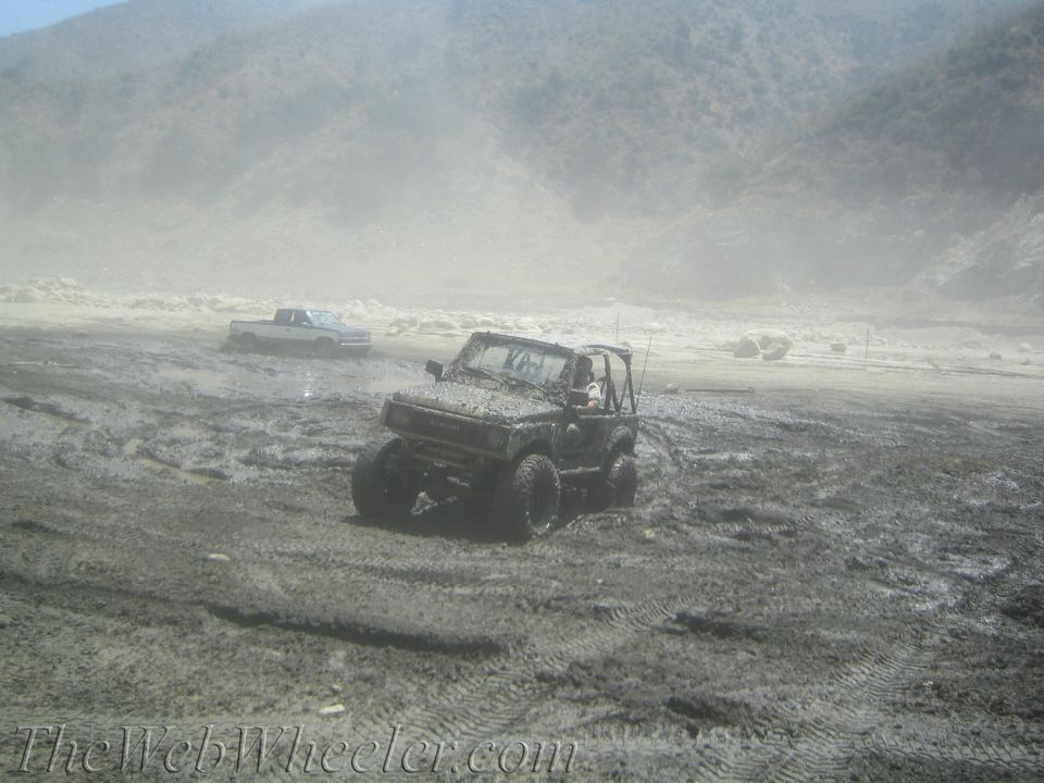 Mud-covered Suzuki Samurai with large tires in a muddy field, a white pickup truck in the background