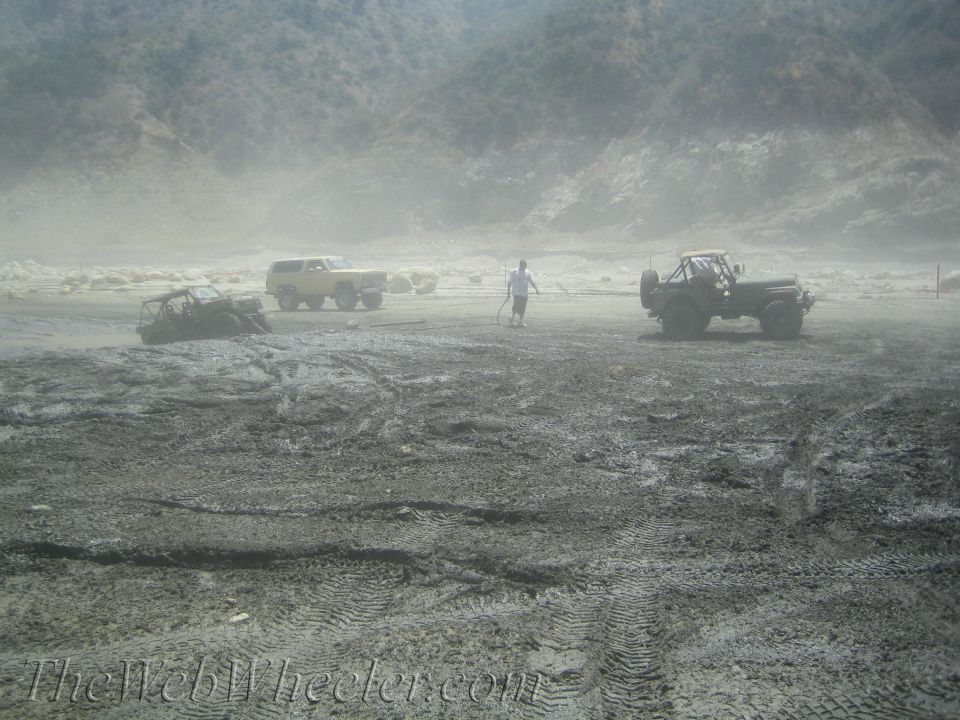 Three off-road vehicles parked in a dusty, muddy area with a person standing between two of them. Hills in the background