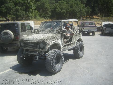1987 Suzuki Samurai - A man sits in a heavily mud-covered off-road vehicle, parked next to other vehicles.