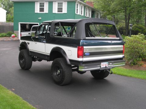 1994 Ford Bronco - A lifted, two-tone Ford Bronco with a soft top is parked on a driveway in front of a green house.
