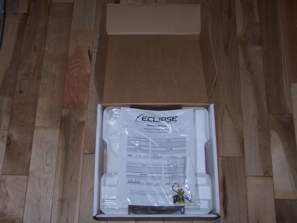 Open box containing an Eclipse car stereo owner's manual and accessories on a wooden floor