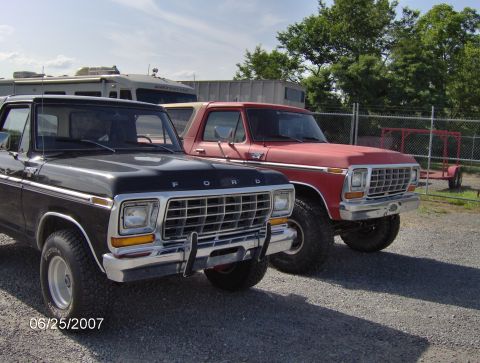 1978 Ford Bronco - Two Ford Bronco trucks, one black and one red, are parked in a gravel lot. The front grille of both vehicles is visible.