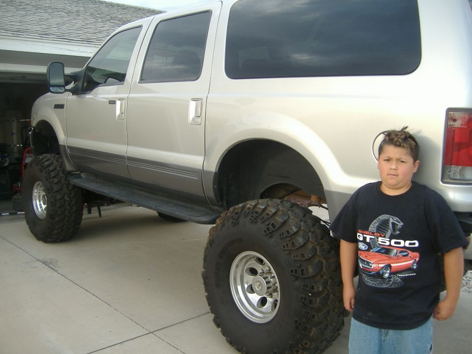 Lifted silver Ford Excursion on huge off-road tires in a driveway with a boy standing beside the rear wheel