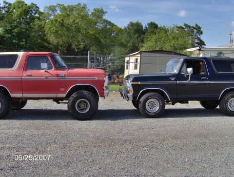 1978 Ford Bronco - Two Ford Broncos, a red and white model and a black model, are shown parked next to each other on a gravel lot.