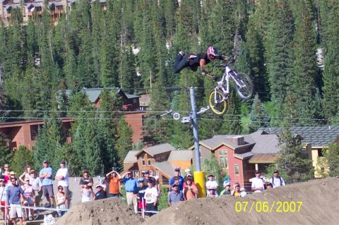 1978 Ford Bronco - A mountain biker performs an aerial stunt over a dirt ramp, with spectators watching and trees in the background.