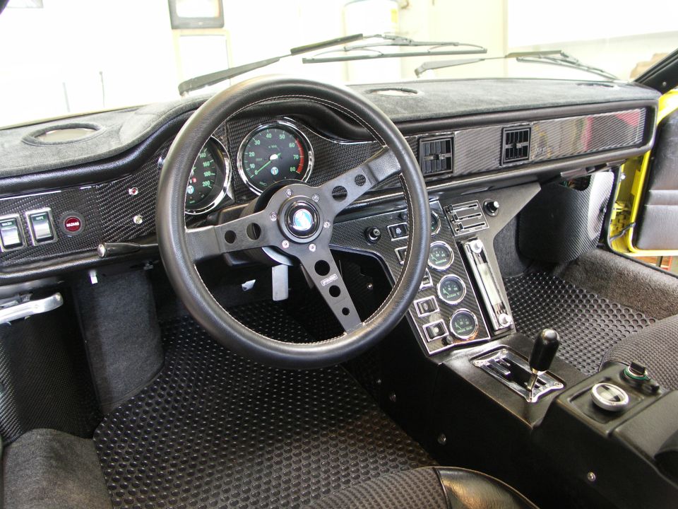 Black interior of a yellow DeTomaso Pantera with carbon-look dash, three-spoke steering wheel, and gated manual shifter