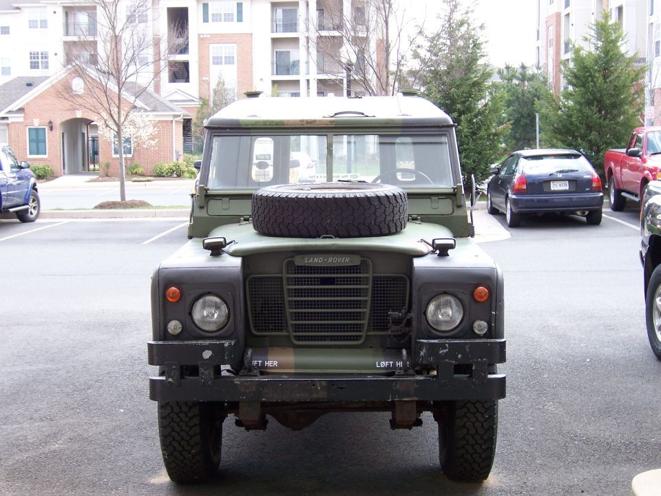 Olive green Land Rover Series III front view with spare tire on the hood, parked in a lot