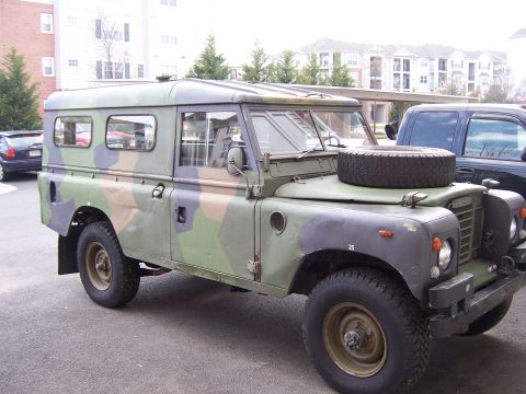 1974 Land Rover Land Rover - A camouflaged Land Rover Defender is parked outdoors, with a dark SUV visible to its right.